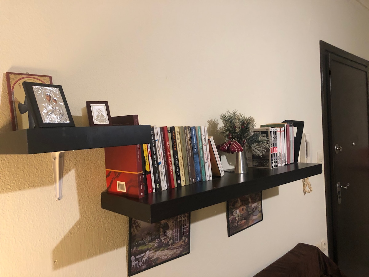 A wall-mounted shelf displays a collection of books alongside framed photographs and a small potted plant. Below the shelf, two framed images are positioned, contributing to the room's character. The minimalist design and organized arrangement create a functional storage solution.