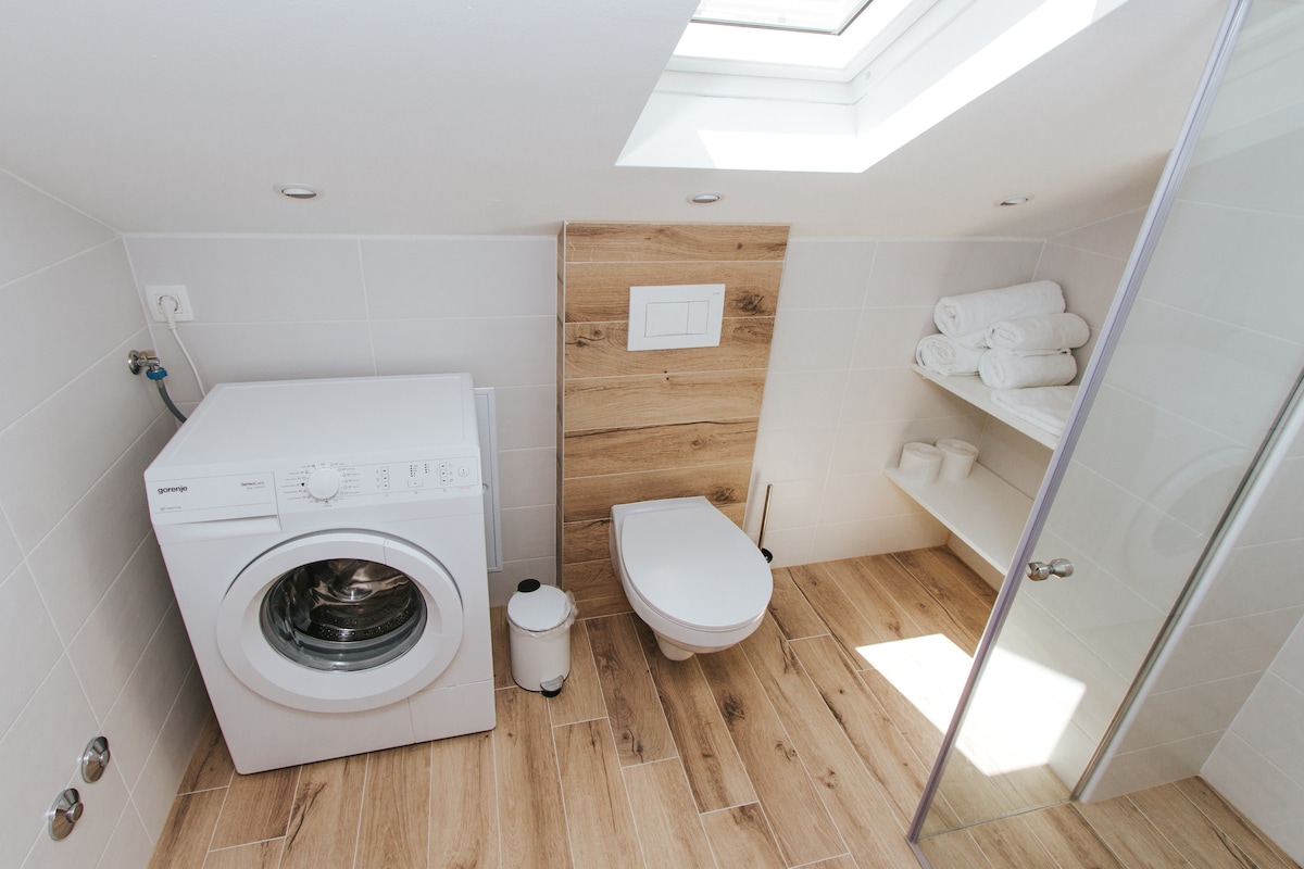 The bathroom features a modern washing machine adjacent to a compact toilet. A sleek shower area is enclosed in glass, and a wooden accent wall adds warmth. Fresh towels are neatly stacked on a wall shelf, enhancing the clean lines of the space, illuminated by natural light from a skylight.