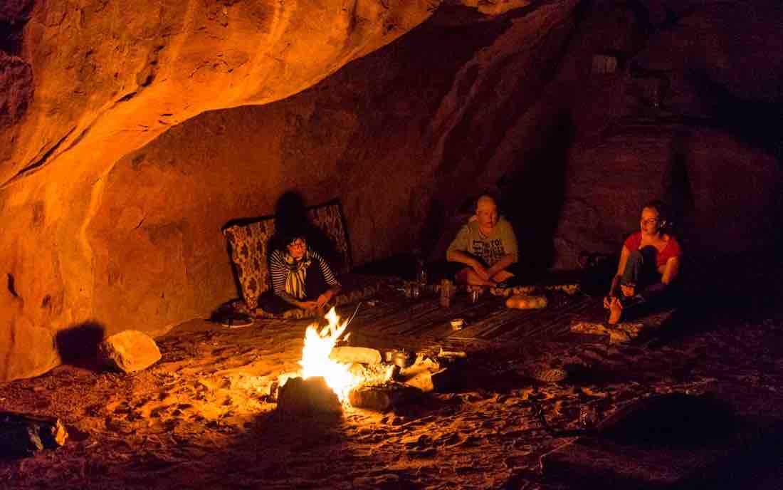A warm fire illuminates a cave setting, where three individuals are seated on woven mats. The rocky walls and floor are visible, and simple food items are arranged nearby, contributing to a cozy atmosphere.