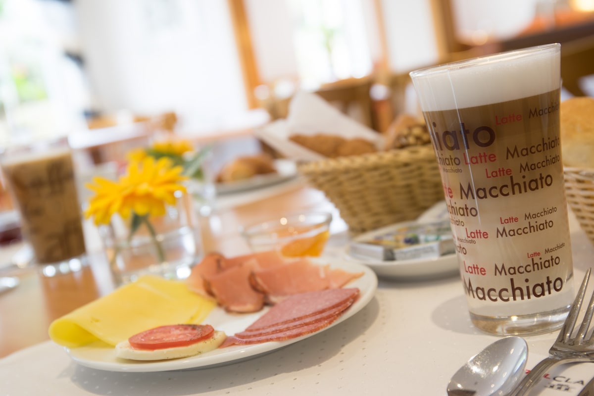 A breakfast setting is presented featuring a glass of latte macchiato and a selection of cold cuts, cheese, and a slice of tomato. A small bowl of jam and a butter dish accompany the meal, with a basket of bread items visible in the background and fresh flowers on the table.