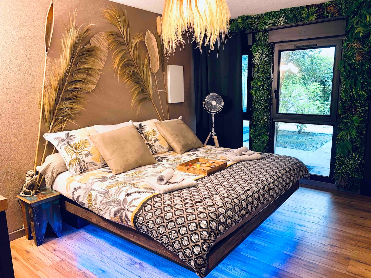 A spacious bed is centered in the room, adorned with decorative cushions and a patterned throw. The walls are complemented by lush greenery and tropical accents, while soft lighting highlights the unique decor. A large window invites natural light and offers views of the outdoor area.