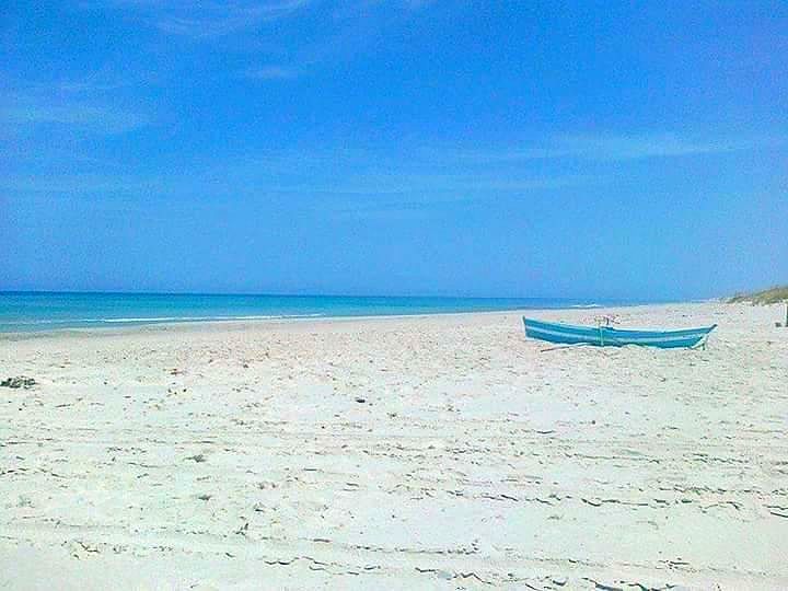 A sandy beach stretches along the shoreline, meeting calm, turquoise waters under a clear blue sky. A small, light blue fishing boat is anchored at the water's edge, highlighting the serene coastal environment.
