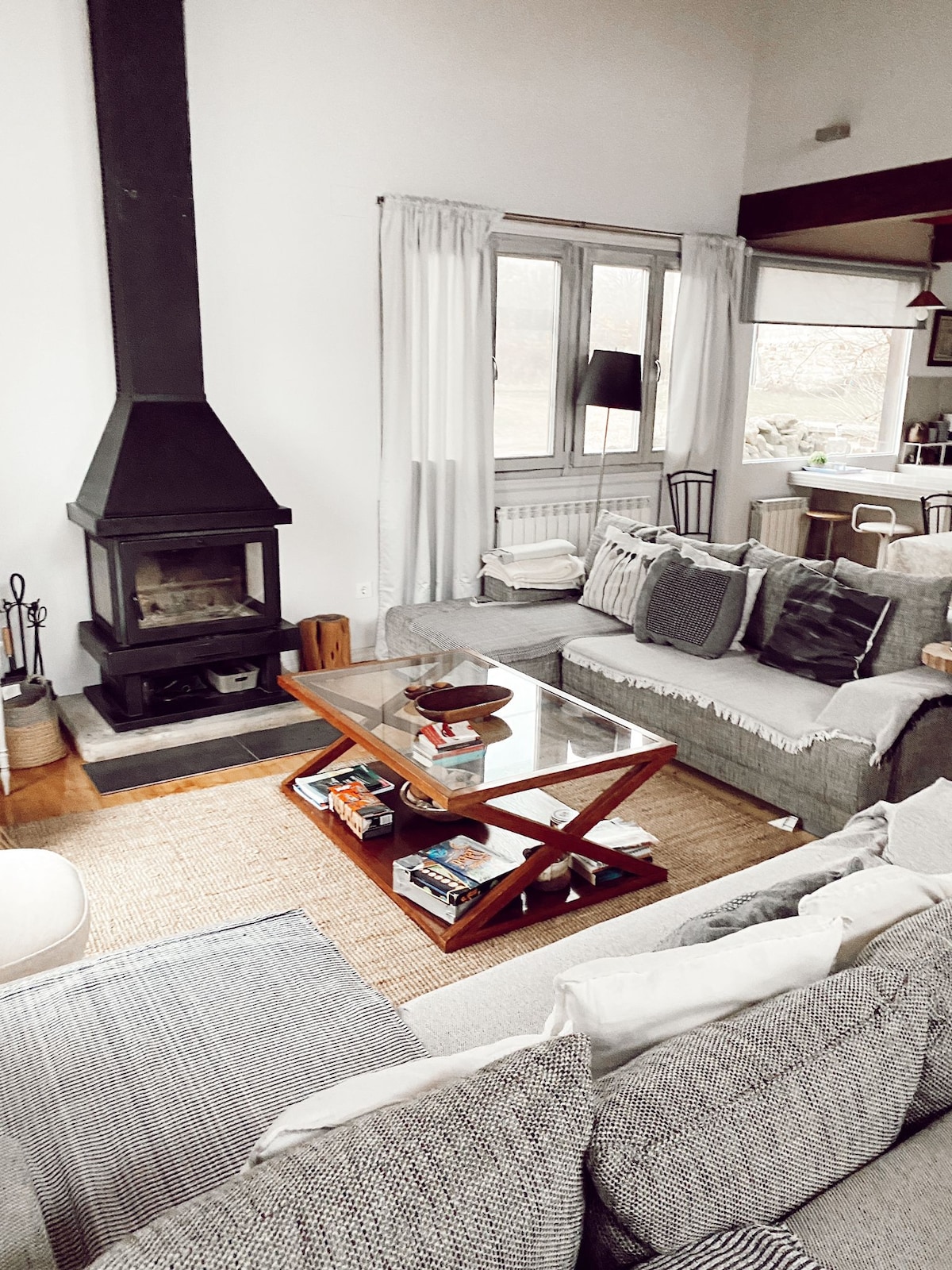 A comfortable living area features a gray sectional sofa and a wooden coffee table. A fireplace is positioned against a wall, while natural light enters through large windows dressed in sheer curtains. Neutral tones create a serene atmosphere, complemented by decorative textiles.