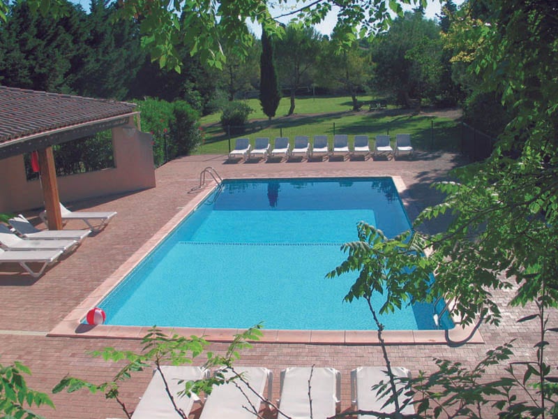 A solar heated swimming pool is surrounded by lounge chairs and a spacious deck. Lush greenery and trees can be seen in the background, providing a sense of privacy and tranquility. Pool floats in red and blue add a touch of color to the inviting scene.