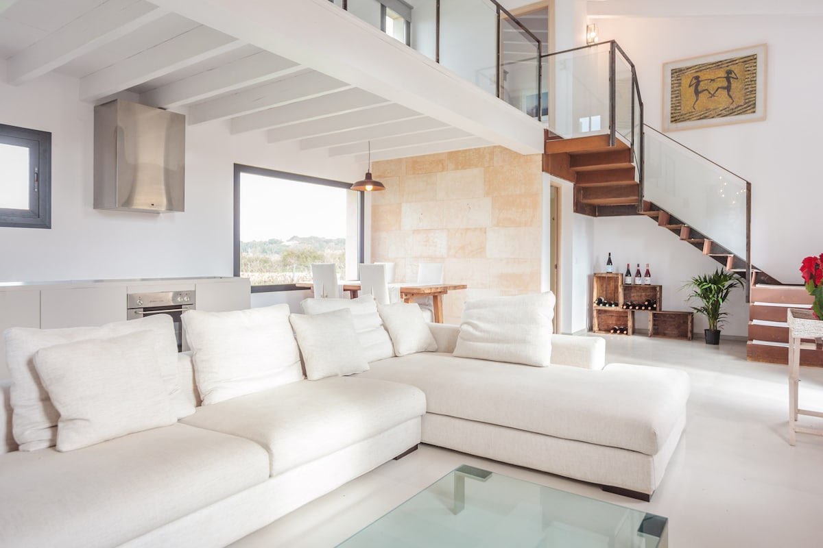 A spacious living area is featured, with a large white sectional sofa positioned around a glass coffee table. Bright light filters through the large windows, illuminating the minimalist design, which includes wooden accents and a staircase leading to an upper level.