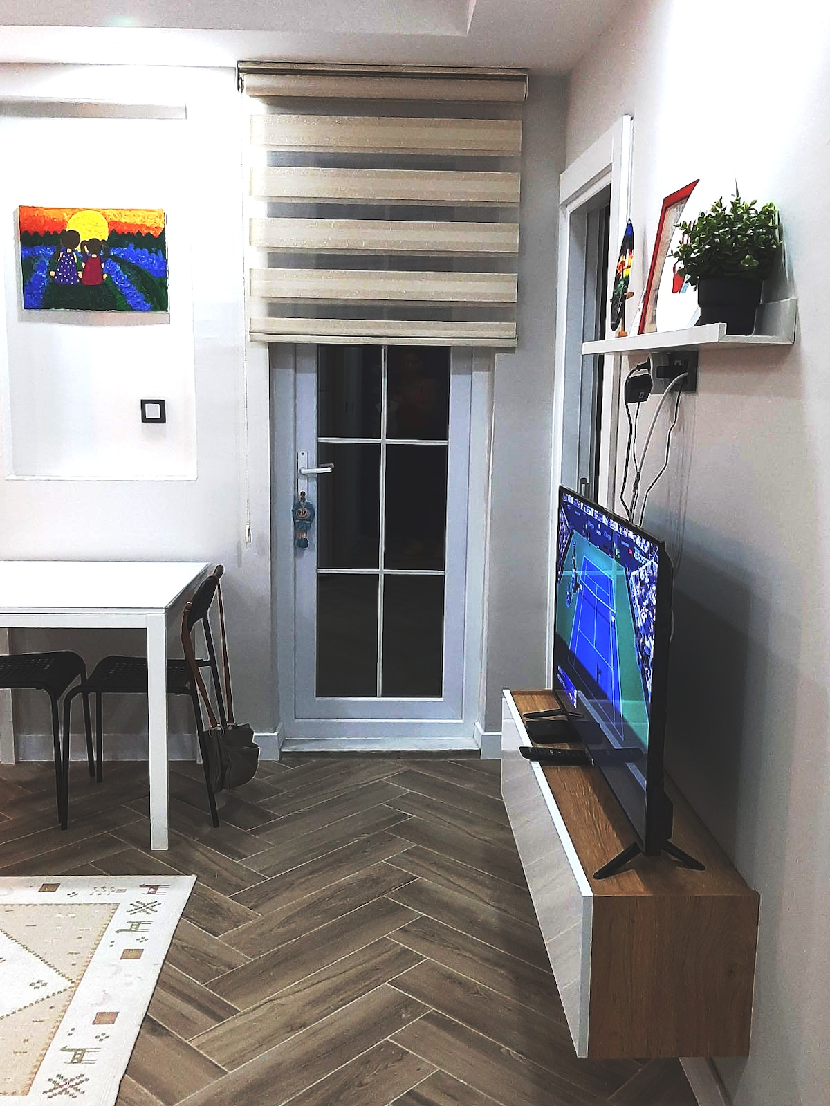 A modern living area is highlighted by a sleek television mounted on a low wooden console. Natural light enters through a glass door adorned with a striped roller shade. A simple dining table is positioned nearby, complemented by black chairs, while colorful artwork decorates the wall.