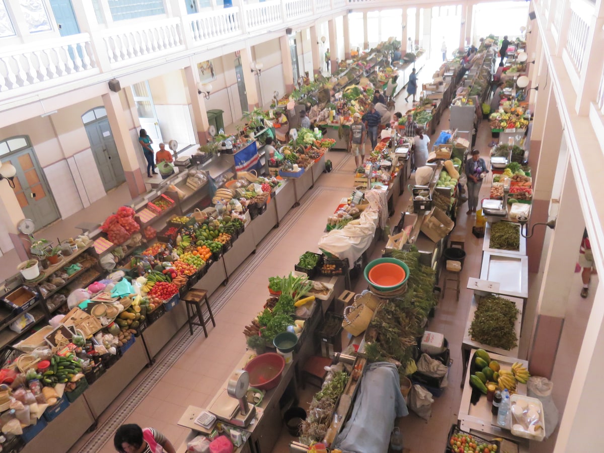 A vibrant market scene displays a variety of fresh produce and goods arranged along long tables. Colorful fruits and vegetables create a lively atmosphere, with shoppers visible throughout the aisles. Natural light filters in from the high windows, illuminating the bustling activity.