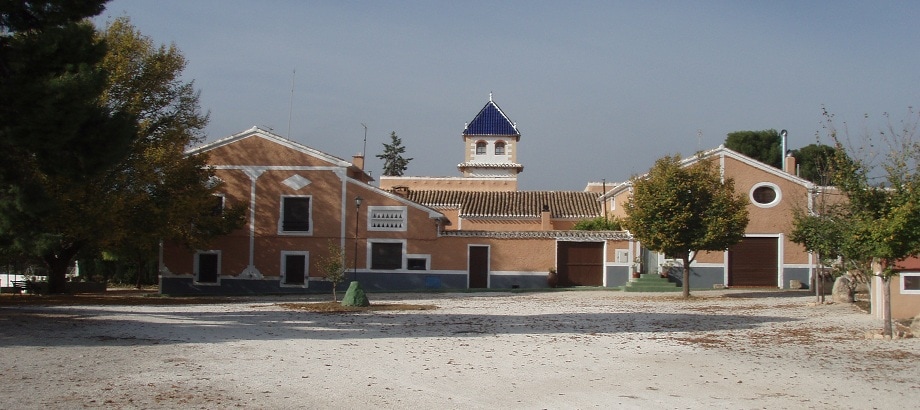 The exterior view of the Cortijo de Rojas showcases a rustic two-story building with a tiled roof and decorative tower. Surrounding trees add greenery to the setting. A gravel driveway leads to the entrance, providing access to the spacious grounds.