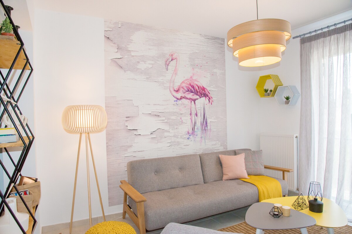 A modern living area features a light grey sofa complemented by a yellow throw and decorative cushions. A striking pink flamingo mural adorns the wall, while ambient lighting is provided by a stylish pendant lamp. Natural light filters through sheer curtains.