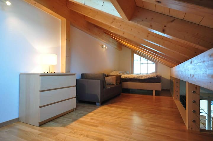 Spavaća soba u galeriji s bračnim krevetom, sofom i stropom do prozora s pogledom na prozor.