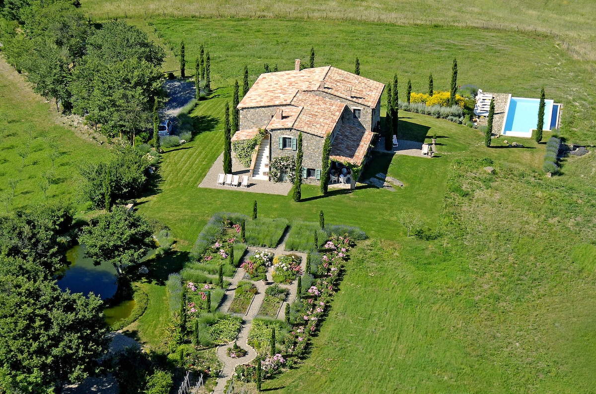 An aerial view captures the stone country house surrounded by lush green landscapes. The house features a terracotta roof and is framed by cypress trees. An infinity pool glistens nearby, while well-maintained gardens and a tranquil pond enhance the natural setting.