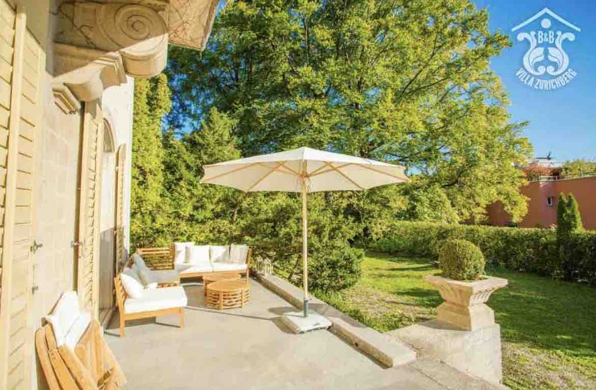 An outdoor patio area is furnished with a white umbrella and light wood seating, featuring plush cushions. Lush greenery surrounds the space, offering a tranquil setting. The patio provides a view of the well-maintained garden and trees, enhancing the overall peaceful ambiance.