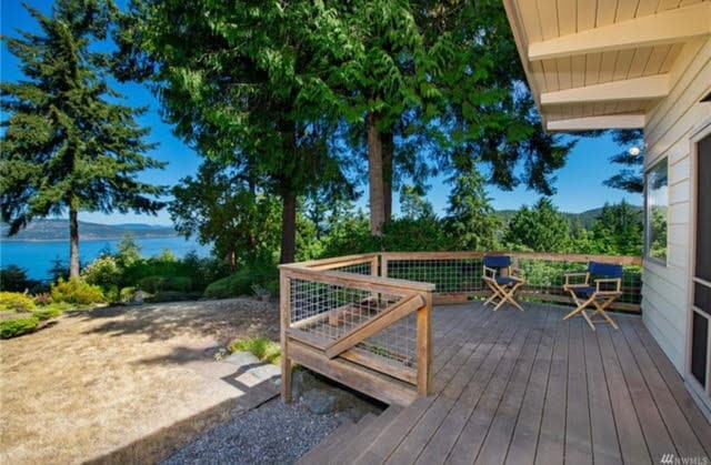 A wooden deck features two blue chairs positioned for optimal views of the surrounding greenery and water. Tall trees provide a natural backdrop, and the lawn leads to a gravel area, enhancing the outdoor space's tranquility.