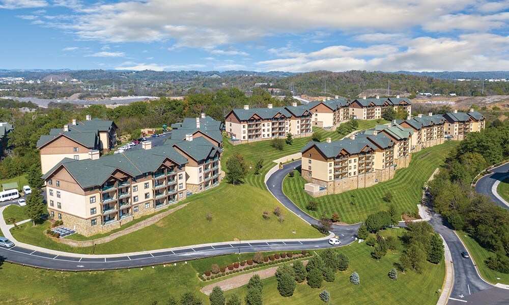 Aerial view of a resort complex featuring multiple multi-story buildings with sloped roofs. Surrounding lawns are well-maintained, showcasing neatly trimmed grass and tree-lined paths. The landscape includes gentle rolling hills, with distant views of mountains under a partly cloudy sky.