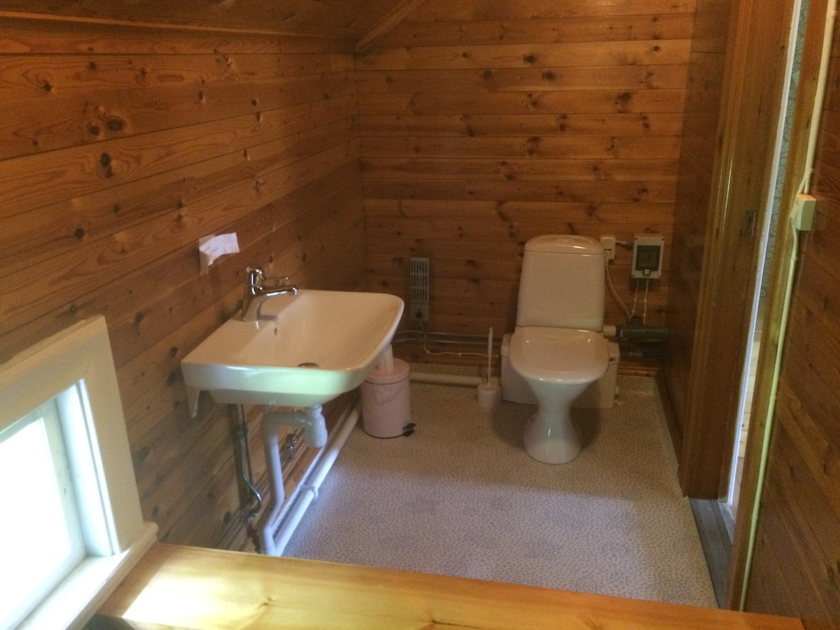 The bathroom features a wooden interior with warm tones, showcasing a white sink and a modern toilet. Natural light enters through a small window, illuminating the space. Basic amenities are neatly arranged, providing a clean and functional environment.