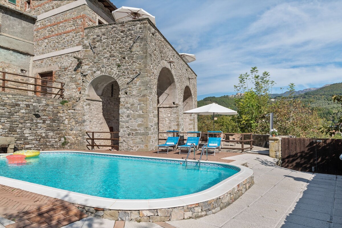 The outdoor area features a shared swimming pool surrounded by sun loungers and parasols. The pool is set against a rustic stone building, complemented by greenery and mountain views in the background. A shaded terrace area provides seating options for relaxation.