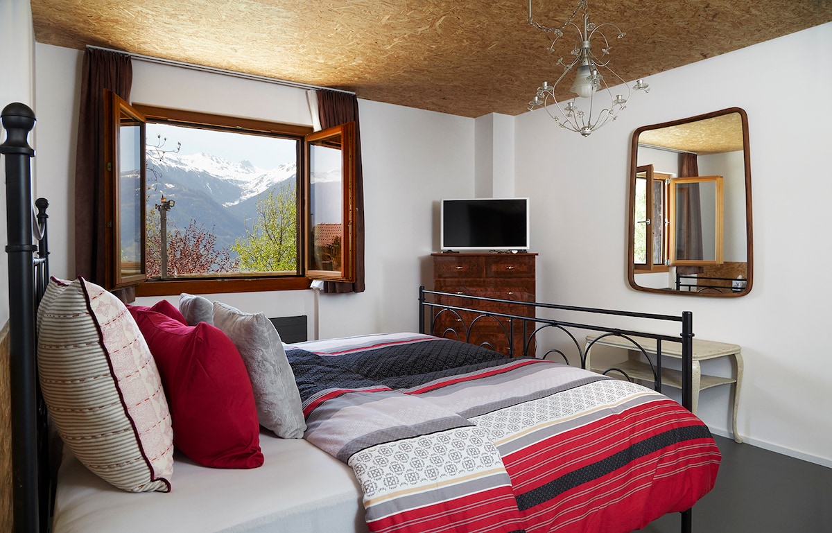 A bright bedroom features a spacious bed adorned with layered bedding in bold patterns. A large window offers a view of the mountains, while soft light filters through. The room includes a mirror, a wooden dresser, and a small table, contributing to a cozy and functional atmosphere.