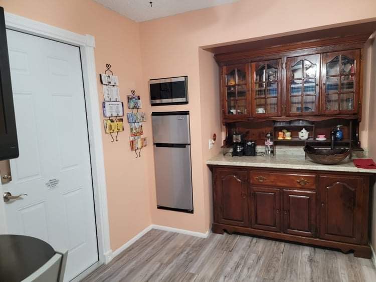 A kitchenette area features wooden cabinetry with glass doors displaying colorful dishes. A sleek stainless refrigerator is set against a light peach wall, and hooks hold various items, including keys and notes. The flooring is light wood, contributing to a bright and functional space.