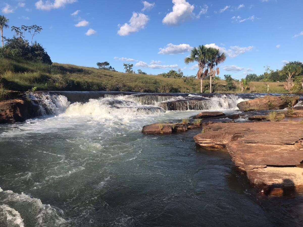 A flowing river cascades over natural rocks, creating a small waterfall. Lush greenery and palm trees surround the scene, with a blue sky adorned with scattered clouds. The tranquil waters are highlighted by sunlight, inviting a sense of peace and connection to nature.