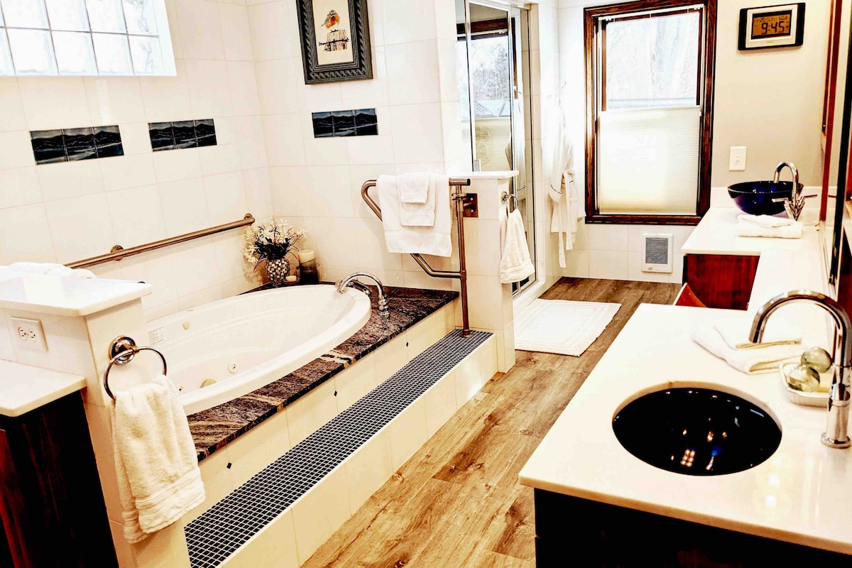 The luxury master bathroom features marble tiled walls, a whirlpool jet tub elevated on a platform, and a glass-enclosed steam shower. Double sinks with sleek design are complemented by modern fixtures. Natural light flows in through multiple windows, enhancing the spacious atmosphere.