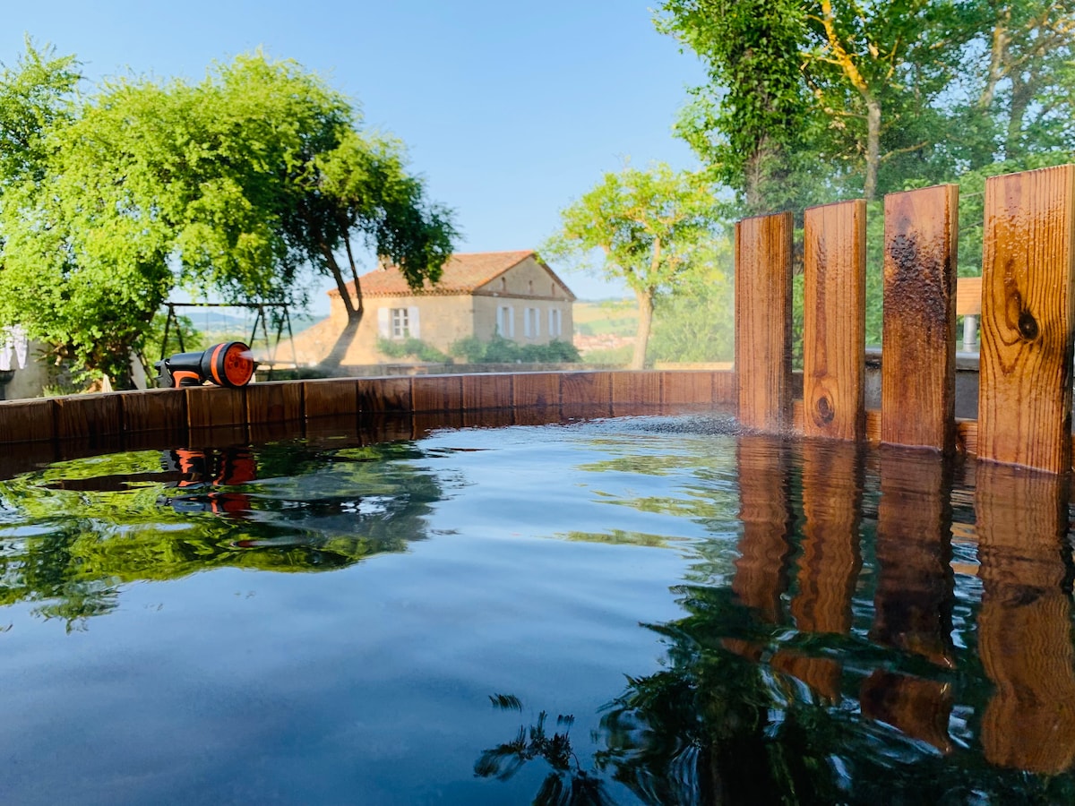 A traditional Finnish hot tub is shown, with clear water reflecting surrounding greenery. The wooden structure surrounds the tub, and a distant house can be seen framed by trees under a clear blue sky.