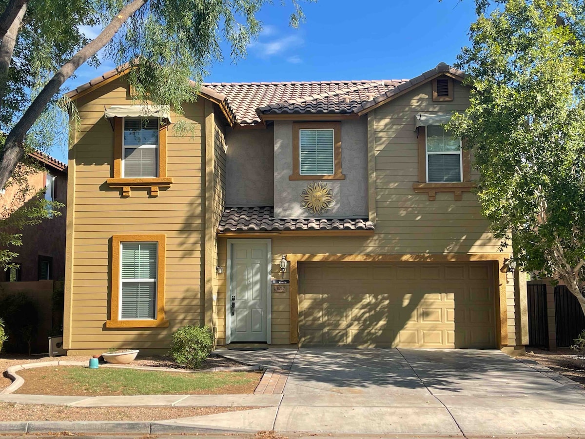 A two-story home with a warm yellow exterior is framed by trees. The front features decorative shutters and a sun emblem above the entrance. A spacious driveway leads to a garage with a taupe door, enhancing the inviting facade.