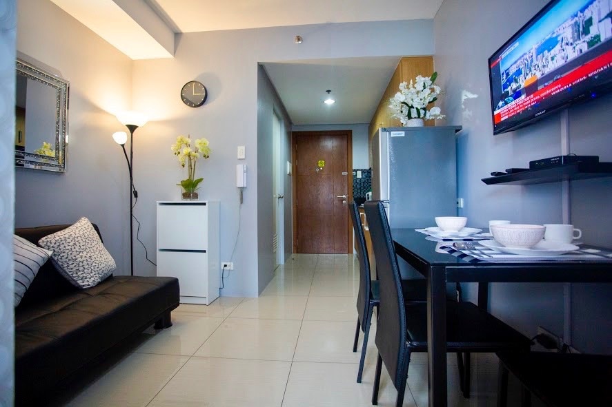 A cozy living area features a sleek black sofa and a dining table, accommodating four chairs. A flat-screen TV is mounted on the wall, and a refrigerator is positioned nearby. The space is illuminated with natural light, creating an inviting atmosphere.