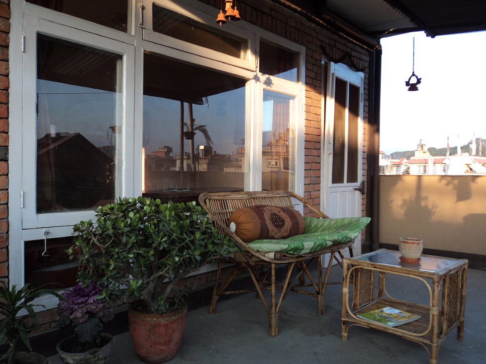 A private terrace garden is featured, showcasing a rattan seating area with decorative pillows. A small table sits nearby, accompanied by potted plants that add greenery. Large windows allow for natural light and views of the surroundings, enhancing the serene outdoor space.