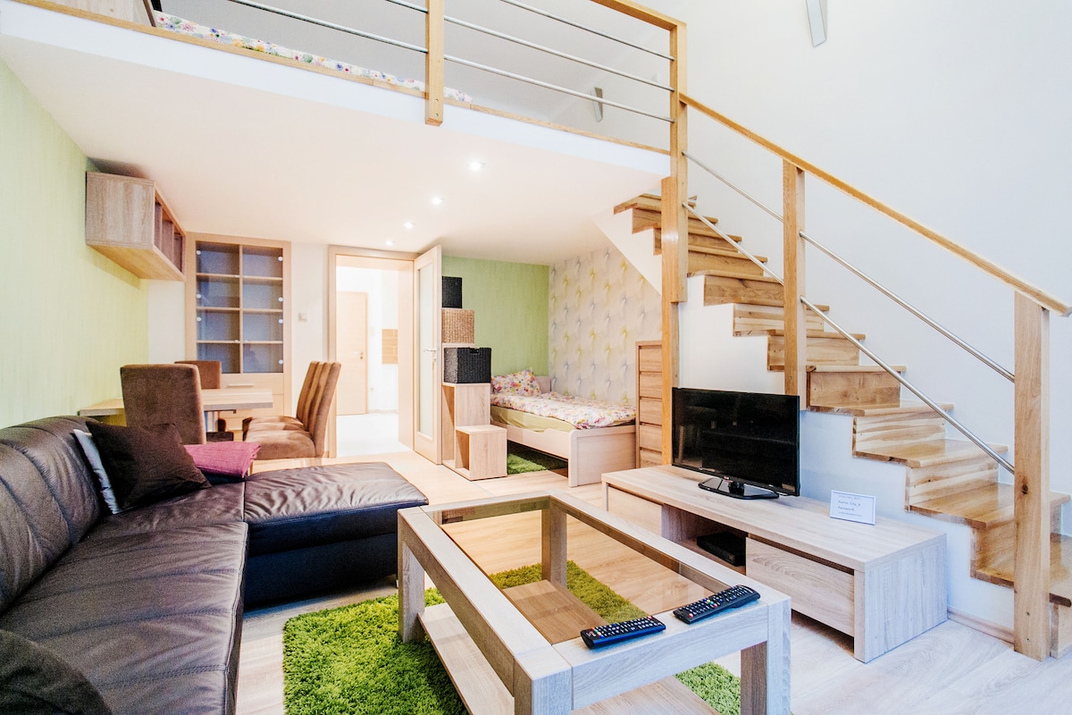 An open living area features a dark leather sectional sofa and a wooden coffee table on a green carpet. A TV is positioned on a light-colored cabinet. Stairs lead to the upper level, where a cozy sleeping area is visible, highlighted by patterned wallpaper.