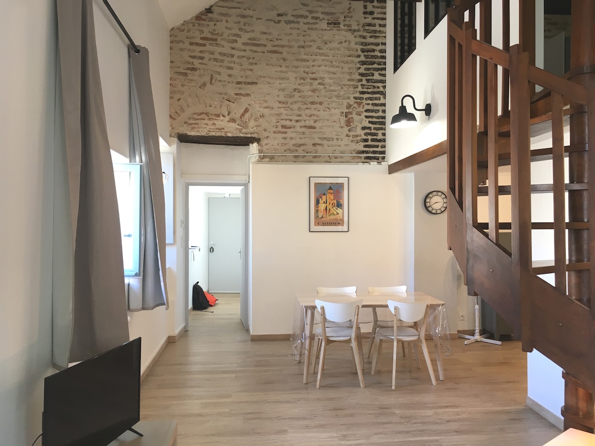 A spacious living area features light wood flooring and exposed brick walls, creating a bright ambiance. A wooden staircase leads to the mezzanine. A dining table with white chairs is positioned near a television, with soft drapes framing the windows.
