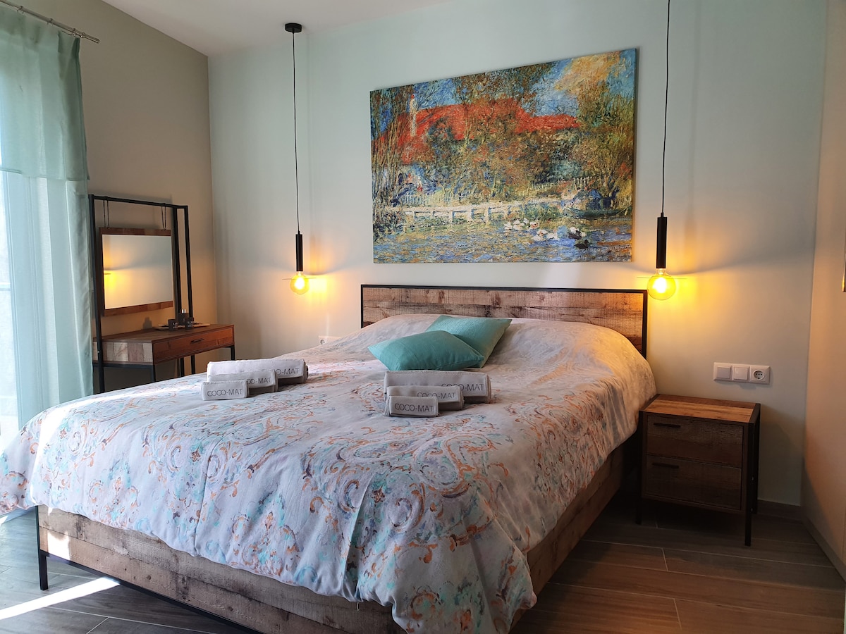 A spacious bedroom features a large bed adorned with a patterned comforter and decorative pillows. Soft pendant lights hang from the ceiling, illuminating the space. A piece of art depicting a colorful landscape is displayed above the bed, enhancing the calming ambiance.
