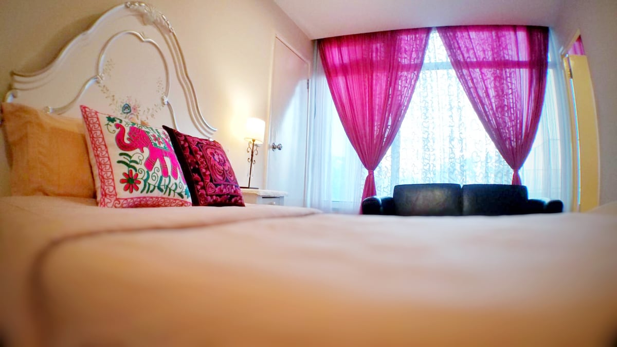 A neatly arranged sleeping area features a spacious bed adorned with colorful decorative pillows. Large windows, dressed in sheer pink curtains, allow natural light to filter through, illuminating the cozy seating area with a dark sofa.