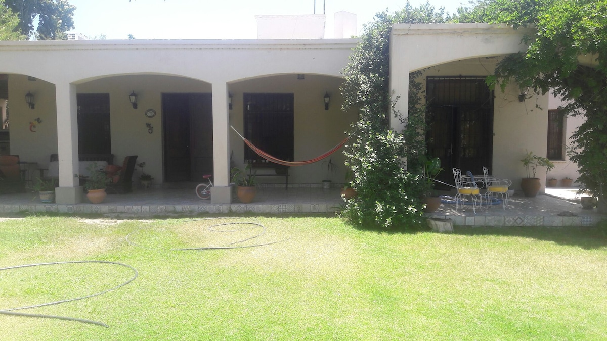 A spacious patio area is depicted, featuring arched columns and potted plants. A hammock is strung between two columns, and a small table with chairs is visible to the right. The well-maintained lawn offers a vibrant green backdrop, enhancing the outdoor ambiance.