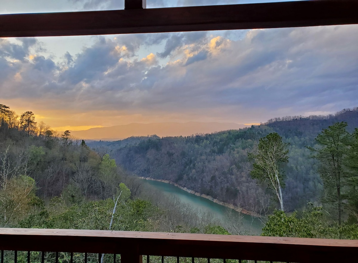 A scenic view from the porch captures a winding river bordered by lush greenery, with distant mountains under a dramatic sky. Warm tones of sunset are visible in the horizon, creating a serene atmosphere.