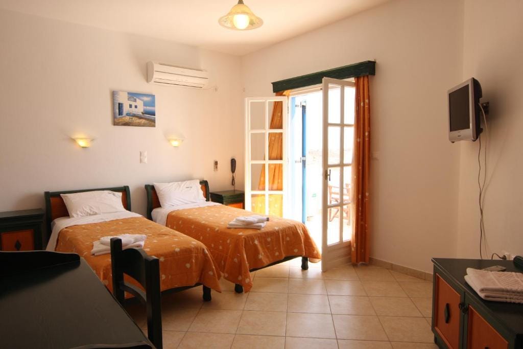 The room features two separate beds draped in bright orange coverings, positioned opposite each other. Natural light floods the space through glass doors leading to a private balcony. A small television is mounted on the wall, and a simple desk arrangement is visible.