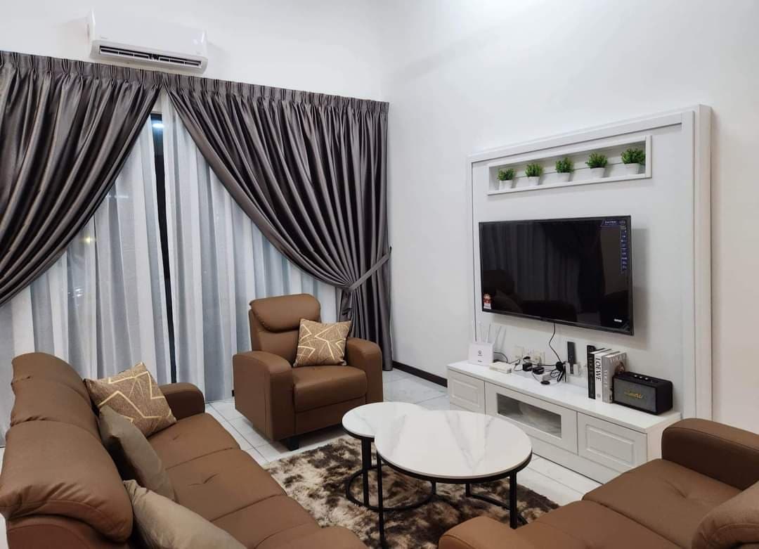 A comfortable living area is showcased with two brown sofas and two armchairs, arranged around a round coffee table. A flat-screen television is mounted on the wall, complemented by decorative plants on a shelf. Floor-to-ceiling curtains provide privacy while allowing natural light.