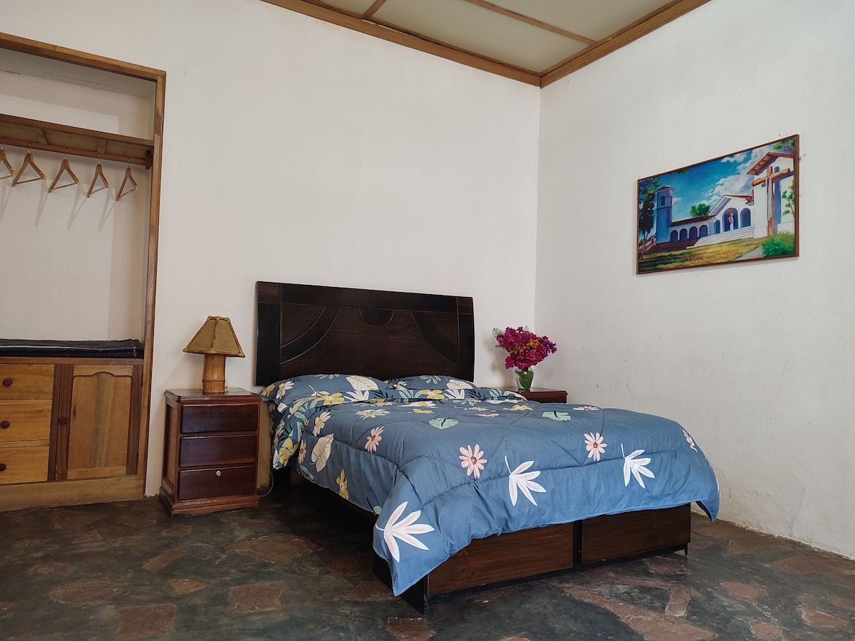 A simple bedroom is presented with a spacious bed adorned in a floral blue duvet. A bedside table and lamp are positioned beside the bed. A colorful painting and a vase of flowers add a touch of decor to the white walls.