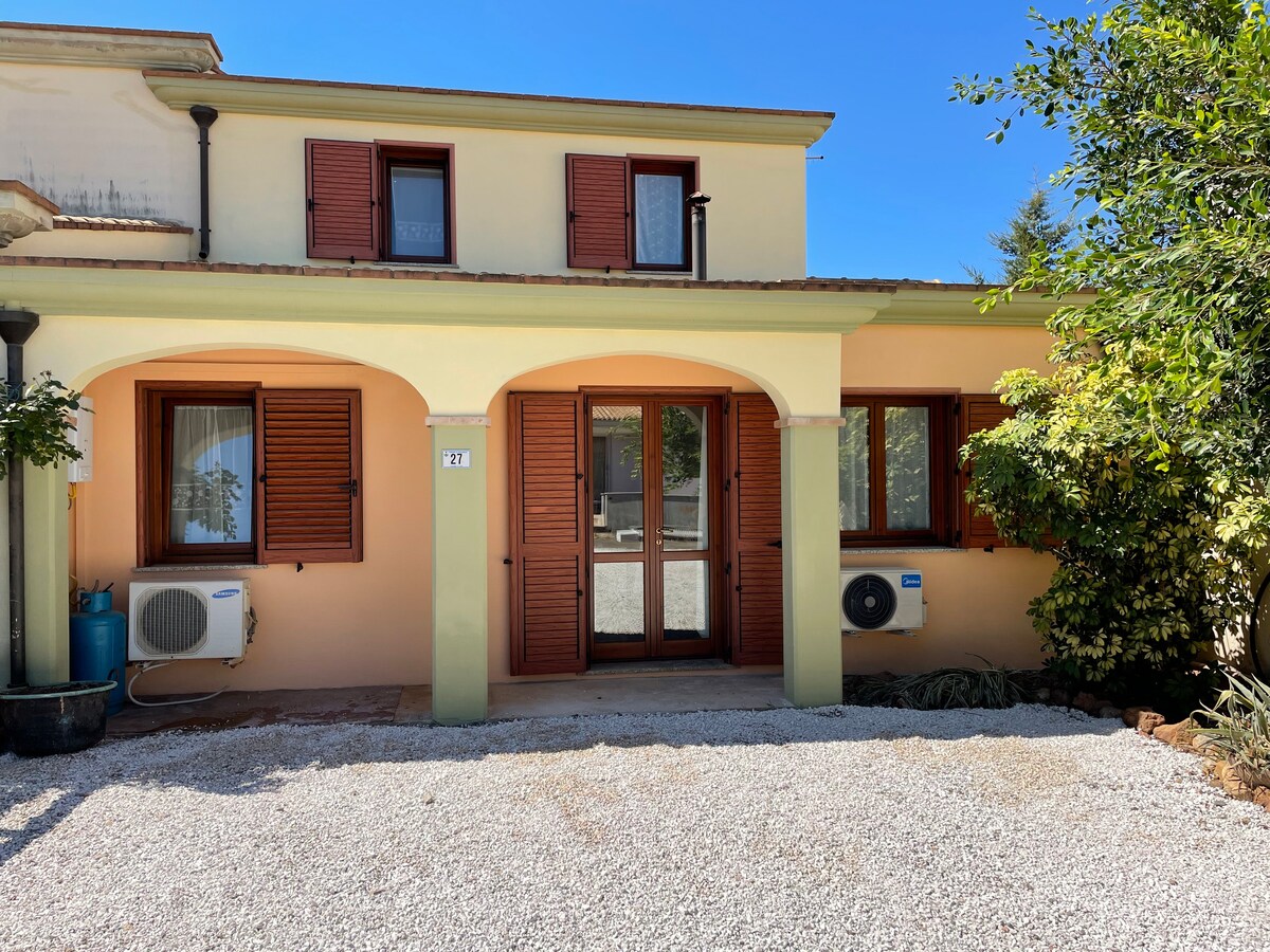 A light yellow building is presented with brown shutters on windows, set against a clear blue sky. A gravel driveway leads to the entrance, framed by greenery and air conditioning units on either side.