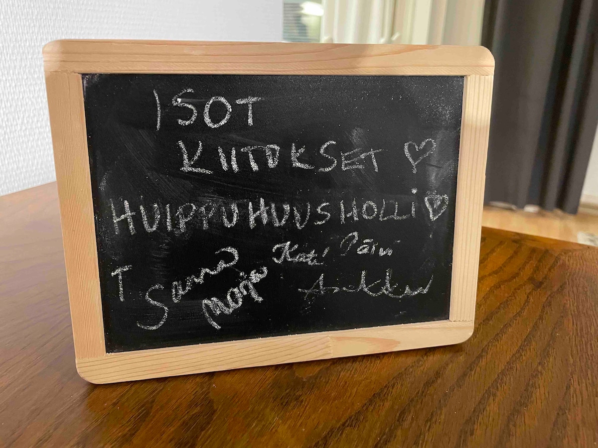 A small wooden chalkboard is displayed on a table, featuring handwritten messages in white chalk. The board's simple design complements the surrounding decor, with a blurred background hinting at a cozy indoor environment.