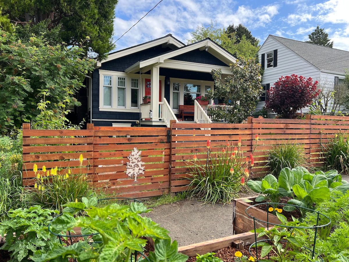 The charming exterior of the home features a welcoming porch with seating, framed by lush green plants and colorful flowers. A wooden fence with decorative cutouts separates the property from the garden area, which displays raised beds filled with various vegetables and vibrant blooms.