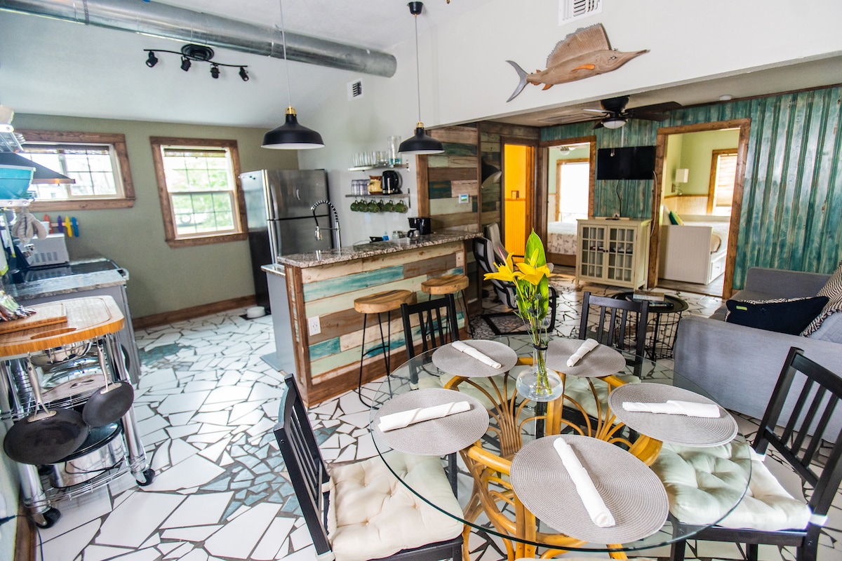 An open-plan living space features a glass dining table surrounded by black wooden chairs. The kitchen is equipped with stainless steel appliances and a breakfast bar. Soft sunlight enters through a window, illuminating the unique patterned tile flooring.