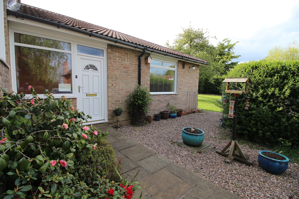 The entrance of the ground floor annex is shown, featuring a welcoming front door framed by neatly manicured gardens. Colorful planters with blooming flowers adorn the path, and a bird feeder stands nearby, enhancing the tranquil outdoor setting.