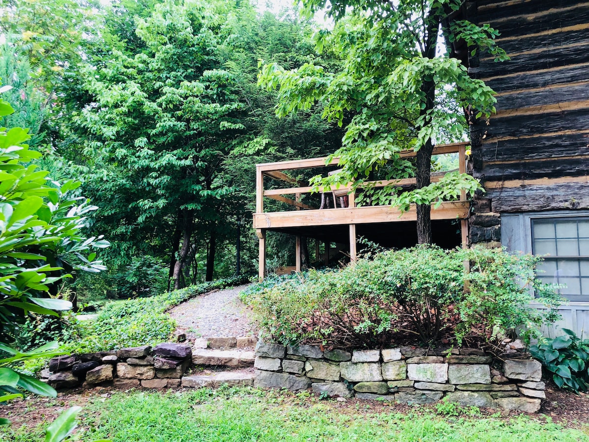 A wooden deck extends from the log cabin, surrounded by lush greenery. The pathway leading up to the deck is bordered by a stone wall, with a gentle incline. Nearby trees provide shade, creating a serene outdoor space perfect for relaxation.