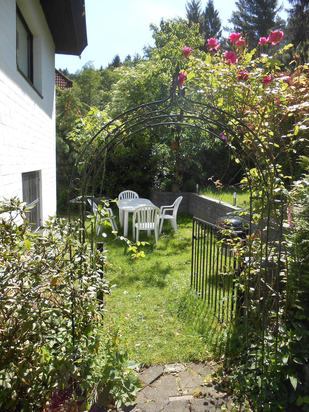 A garden archway adorned with blooming flowers frames a view of a grassy area. In the foreground, a small table is surrounded by four white plastic chairs, offering a serene outdoor seating space. Lush greenery surrounds the setting, contributing to a peaceful ambiance.