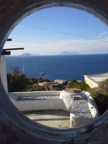 A round opening frames a view of the blue sea and distant islands. The horizon meets the water in the background, with white clouds scattered across a blue sky. A small boat is visible on the water, while greenery and stone walls are seen in the foreground.