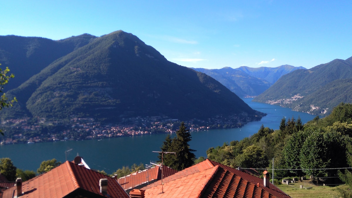 A panoramic view of Lake Como is displayed, with mountains framing the water on either side. The rooftops of charming houses are visible in the foreground, while the lush landscape and the calm lake reflect the bright blue sky.