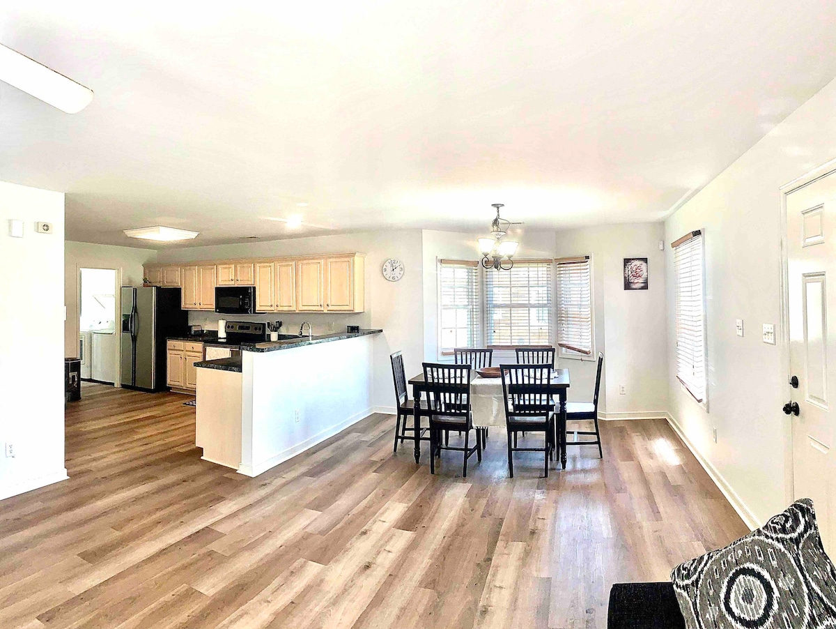 An open living and dining area is highlighted by a wooden dining table surrounded by six chairs. The kitchen features light cabinetry and modern appliances. Natural light floods the space through large windows, and a cozy couch is present in the foreground.