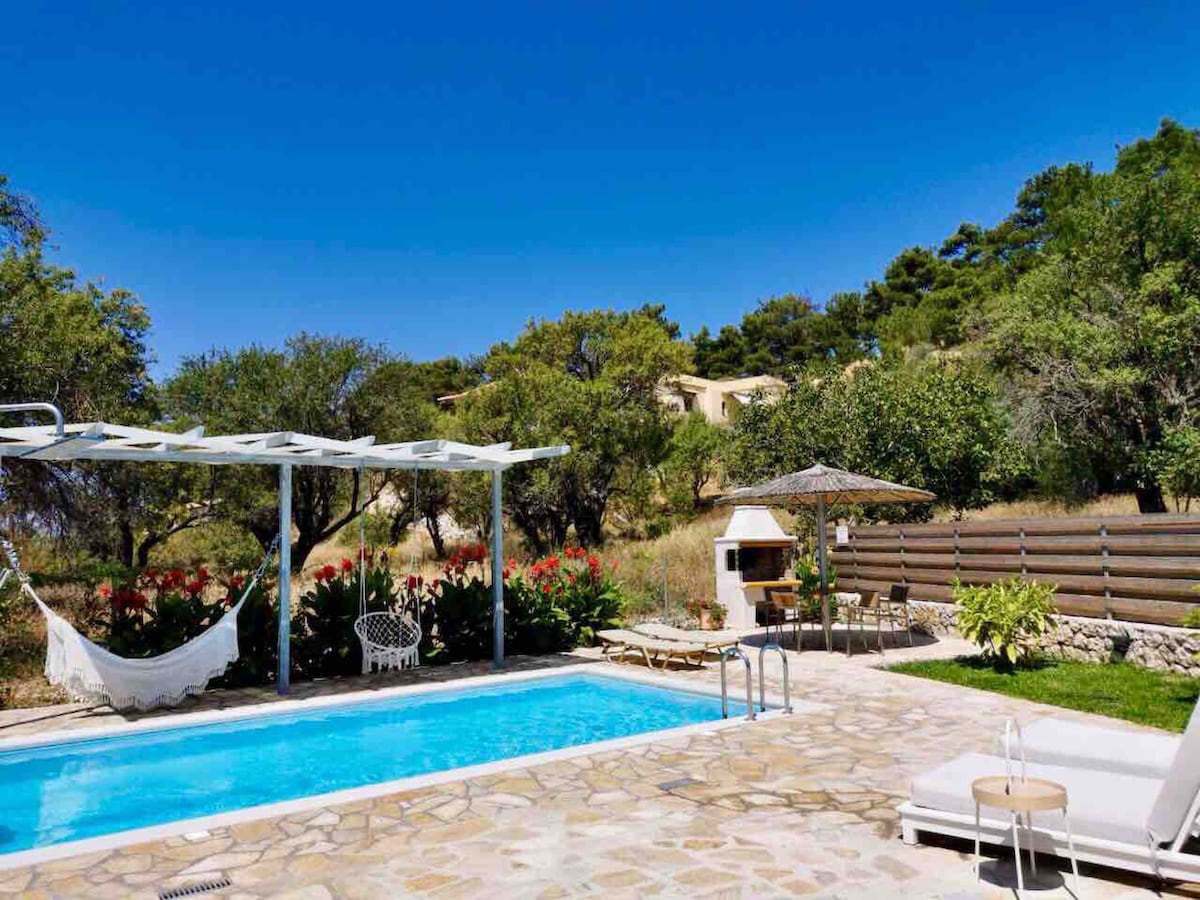 A private saltwater pool area is surrounded by lush greenery, featuring large sunbeds and an outdoor shower. A BBQ area and a shaded sitting space with garden furniture are visible, providing a relaxing atmosphere for outdoor dining. The clear blue sky enhances the serene setting.