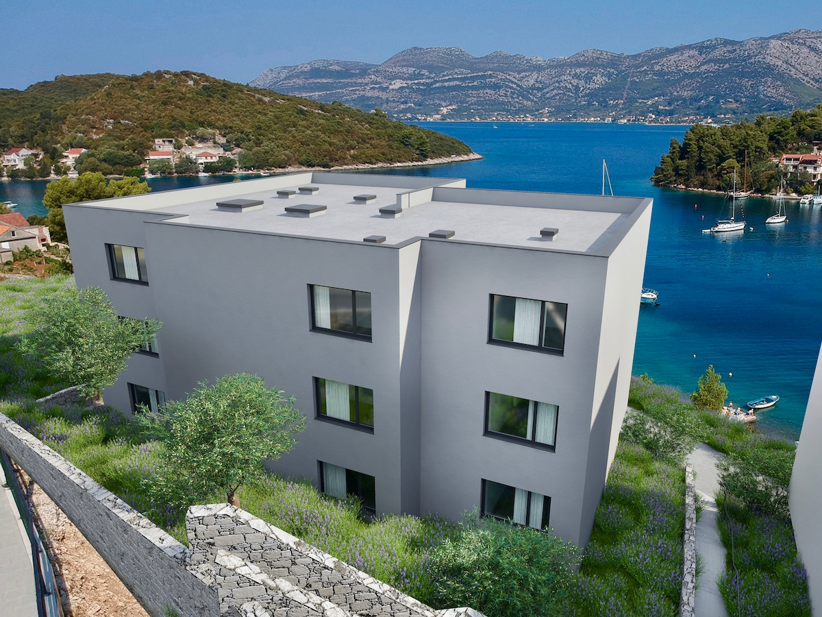 A modern apartment complex is set against a backdrop of the calm blue bay, surrounded by lush greenery. The building features multiple large windows that invite natural light, while the serene water reflects the picturesque landscape of nearby hills and boats.