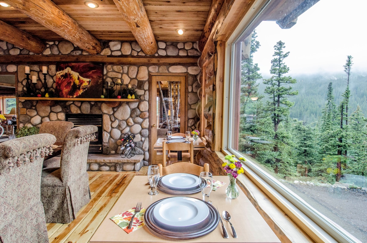 A dining area features wood beams and a stone fireplace, with two elegantly set tables near expansive windows. The view outside includes tall trees and distant mountains, while flowers add a touch of color to the table settings.